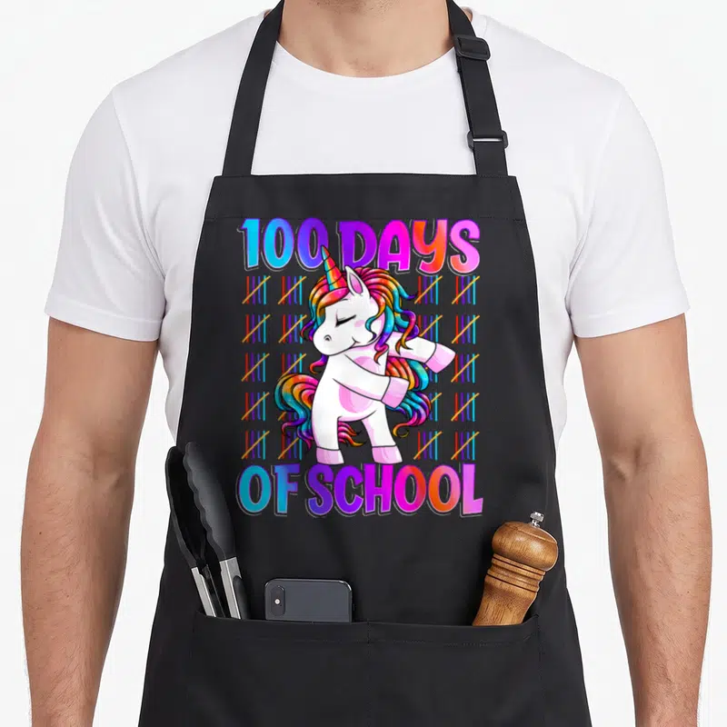 Smarter unicorn apron - Cartoon unicorn dabbing with 100 DAYS OF SCHOOL text and rainbow tally marks — shown on a black cotton canvas apron against a white background