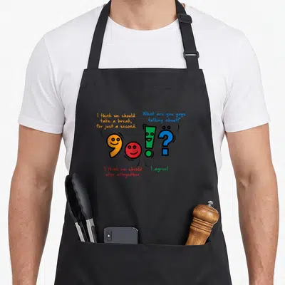 Punctuation english apron - Four colorful punctuation marks with faces and speech bubbles arranged in a row — shown on a black cotton canvas apron against a white background