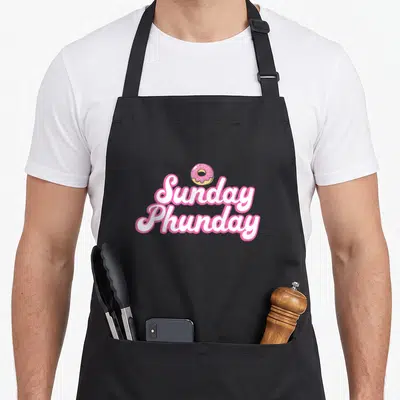 Sunday donut apron - Pink frosted donut above Sunday Phunday white script text with pink outline — shown on a black cotton canvas apron against a white background