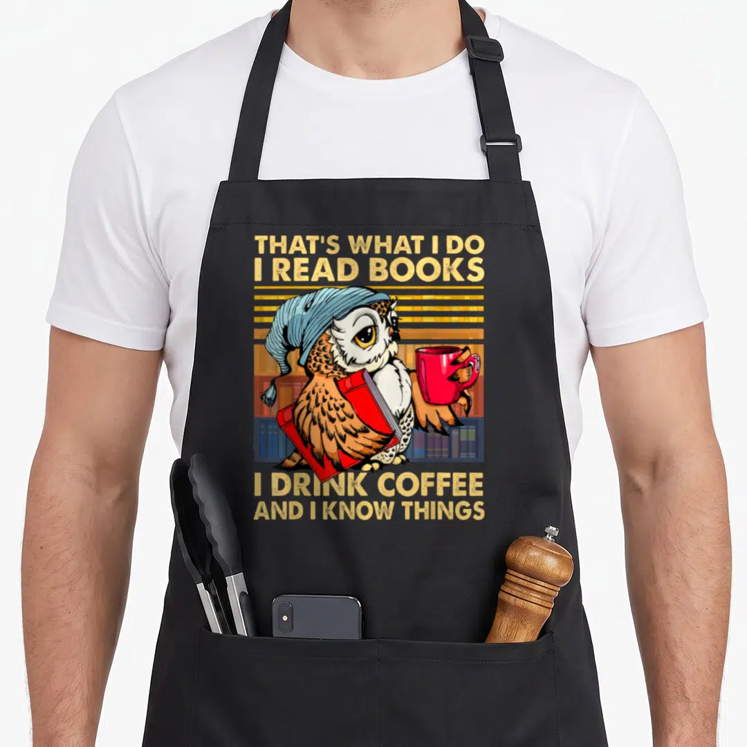 Owl coffee apron - Cartoon owl in blue beanie holding red mug and book with text That's What I Do I Read Books I Drink — shown on a black cotton canvas apron against a white background