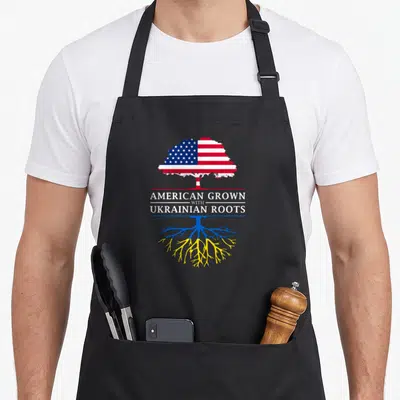 Grown ukrainian aprons - Tree with US flag canopy and Ukrainian colored roots sandwiching text AMERICAN GROWN — shown on a black cotton canvas apron against a white background
