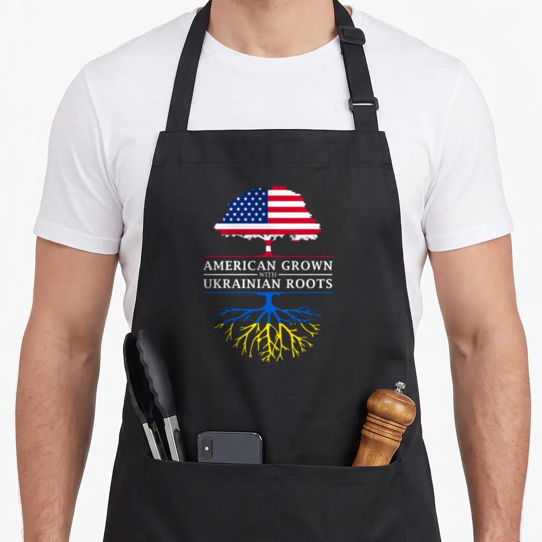 Grown ukrainian aprons - Tree with US flag canopy and Ukrainian colored roots sandwiching text AMERICAN GROWN — shown on a black cotton canvas apron against a white background