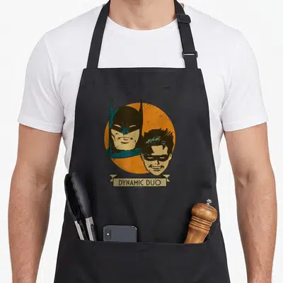 Dynamic animal apron - Batman and Robin heads on orange circle with beige DYNAMIC DUO banner — shown on a black cotton canvas apron against a white background