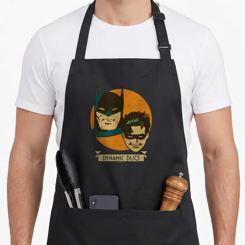 Dynamic animal apron - Batman and Robin heads on orange circle with beige DYNAMIC DUO banner — shown on a black cotton canvas apron against a white background