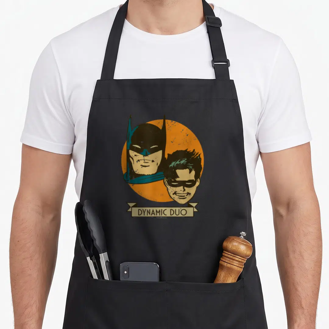 Dynamic animal apron - Batman and Robin heads on orange circle with beige DYNAMIC DUO banner — shown on a black cotton canvas apron against a white background