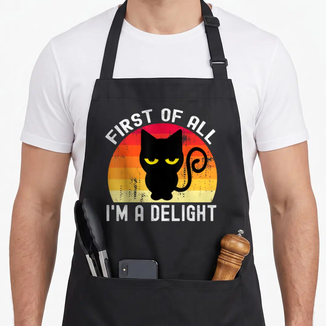 Cat snarky aprons - Black cat silhouette in front of striped sun with white text FIRST OF ALL I'M A DELIGHT — shown on a black cotton canvas apron against a white background