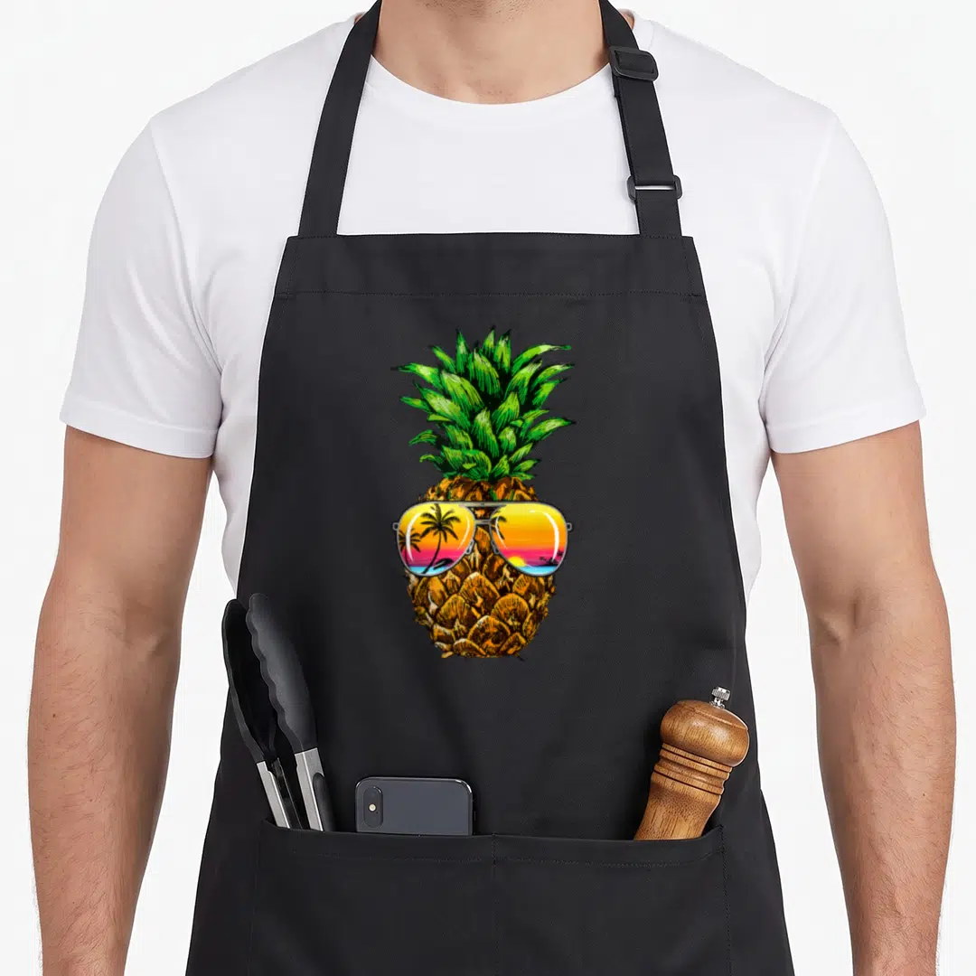 Sunglass hawaiian apron - Cartoon pineapple wearing sunglasses reflecting a palm tree sunset in yellow, green — shown on a black cotton canvas apron against a white background