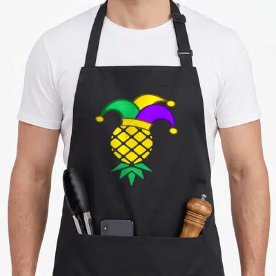 Upside fat chef apron - Inverted yellow pineapple wearing a purple green and yellow jester hat — shown on a black cotton canvas apron against a white background
