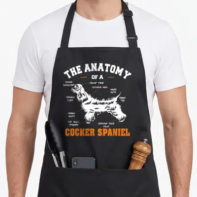 Anatomy cocker spaniel apron - White Cocker Spaniel silhouette with orange labels pointing to soulful eyes and booster — shown on a black cotton canvas apron against a white background