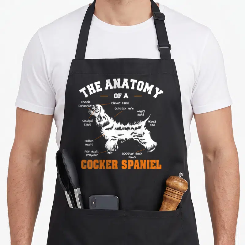 Anatomy cocker spaniel apron - White Cocker Spaniel silhouette with orange labels pointing to soulful eyes and booster — shown on a black cotton canvas apron against a white background