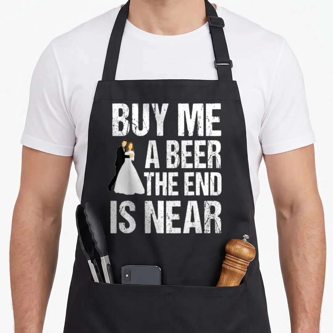 Funny bachelor party apron - White text reading Buy Me A Beer The End Is Near beside cartoon bride and groom — shown on a black cotton canvas apron against a white background