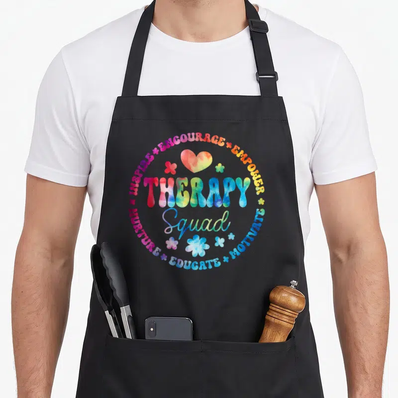 Week my therapy apron - Tie-dye text circle with inspire encourage empower motivate educate nurture therapy squad words — shown on a black cotton canvas apron against a white background
