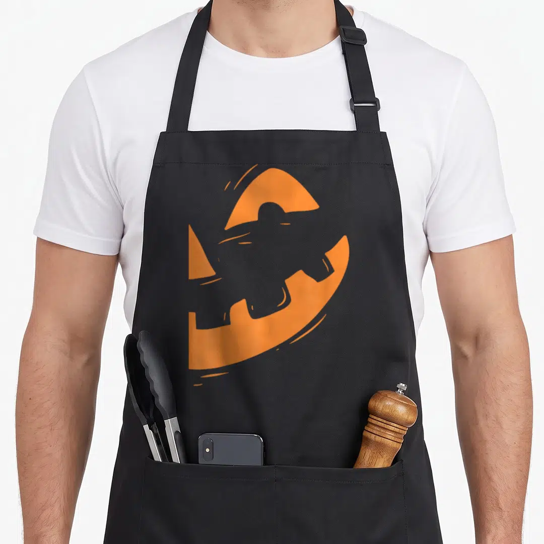 Jack lantern pumpkin halloween apron - Minimalist orange pumpkin with grey jet silhouette negative space design — shown on a black cotton canvas apron against a white background
