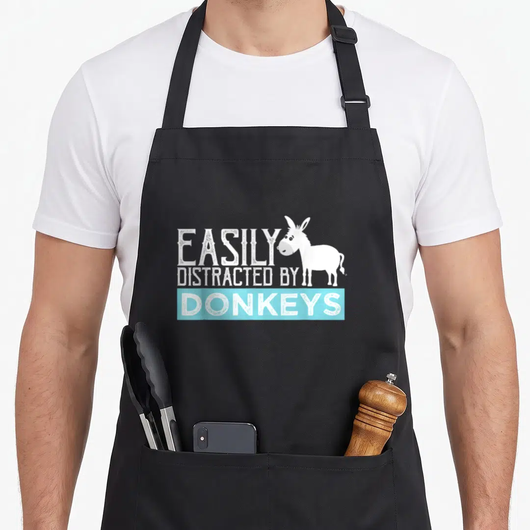 Shirtside donkey apron - Cartoon white donkey beside text easily distracted by donkeys in blue banner — shown on a black cotton canvas apron against a white background