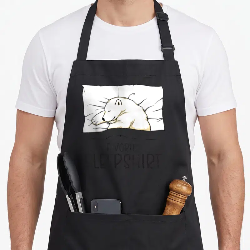Sleepshirt bear apron - Cartoon polar bear lying on white pillow above black text reading FAVORITE SLEEPSHIRT — shown on a black cotton canvas apron against a white background