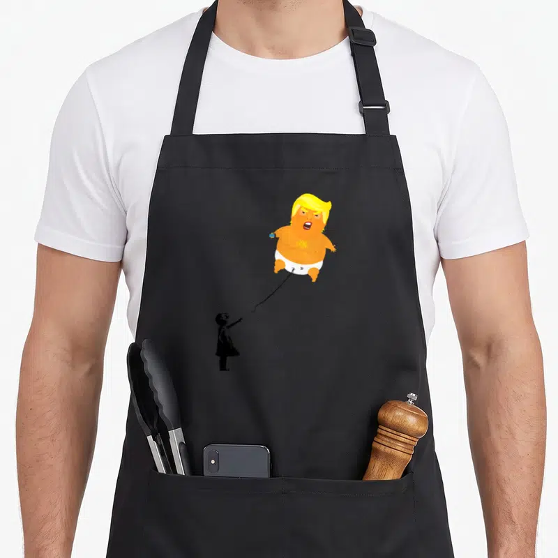 Trump blimp apron - Orange cartoon baby balloon on a leash held by a black child silhouette — shown on a black cotton canvas apron against a white background