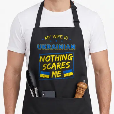 Ukrainian wife apron - Stacked text reading MY WIFE IS UKRAINIAN NOTHING SCARES ME with three stars and two blue yellow — shown on a black cotton canvas apron against a white background