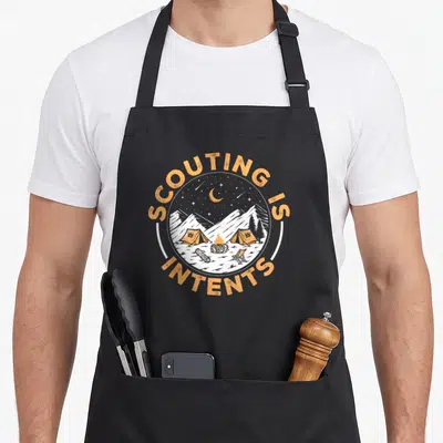Scouting camping apron - Circular campsite with orange tents and fire, surrounded by arched text SCOUTING IS INTENTS — shown on a black cotton canvas apron against a white background