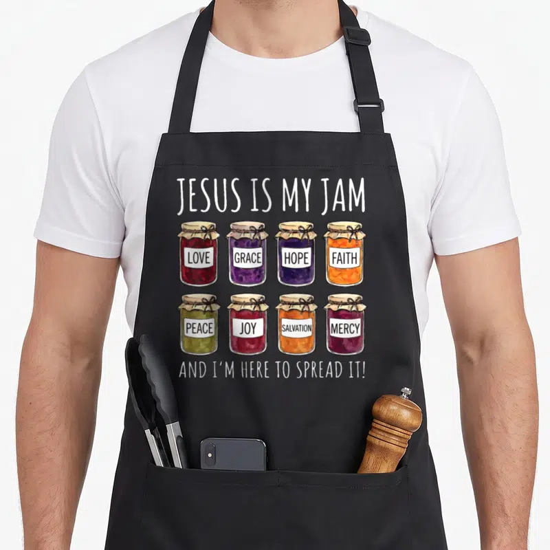 Jesus spread apron - Eight jam jars labeled love grace hope faith peace joy salvation mercy with pun text — shown on a black cotton canvas apron against a white background