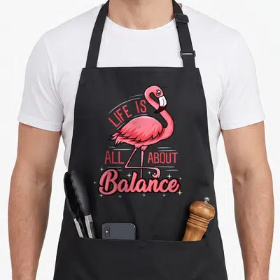 Balance flamingo apron - Pink cartoon flamingo standing on one leg with LIFE IS ALL ABOUT Balance text and stars — shown on a black cotton canvas apron against a white background