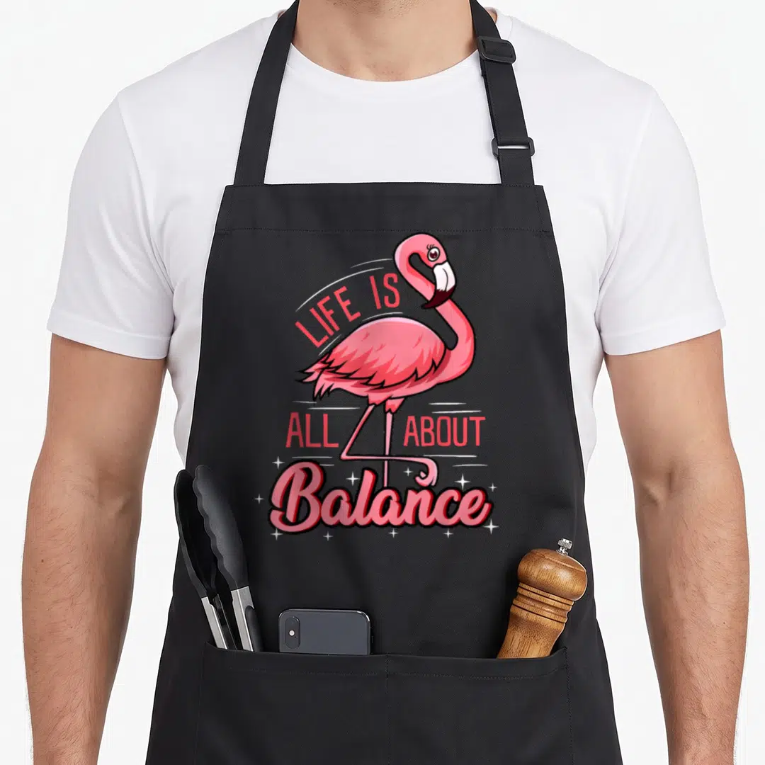 Balance flamingo apron - Pink cartoon flamingo standing on one leg with LIFE IS ALL ABOUT Balance text and stars — shown on a black cotton canvas apron against a white background