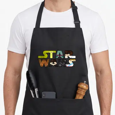 Character kawaii apron - Star Wars text filled with cartoon character faces in green, teal, brown, black, and white — shown on a black cotton canvas apron against a white background