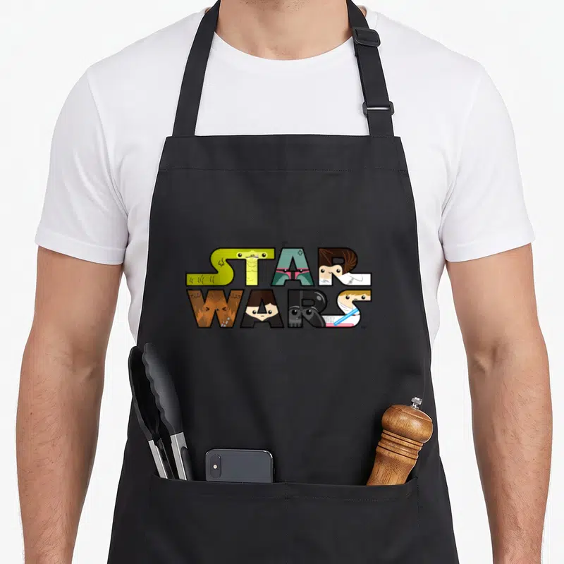 Character kawaii apron - Star Wars text filled with cartoon character faces in green, teal, brown, black, and white — shown on a black cotton canvas apron against a white background