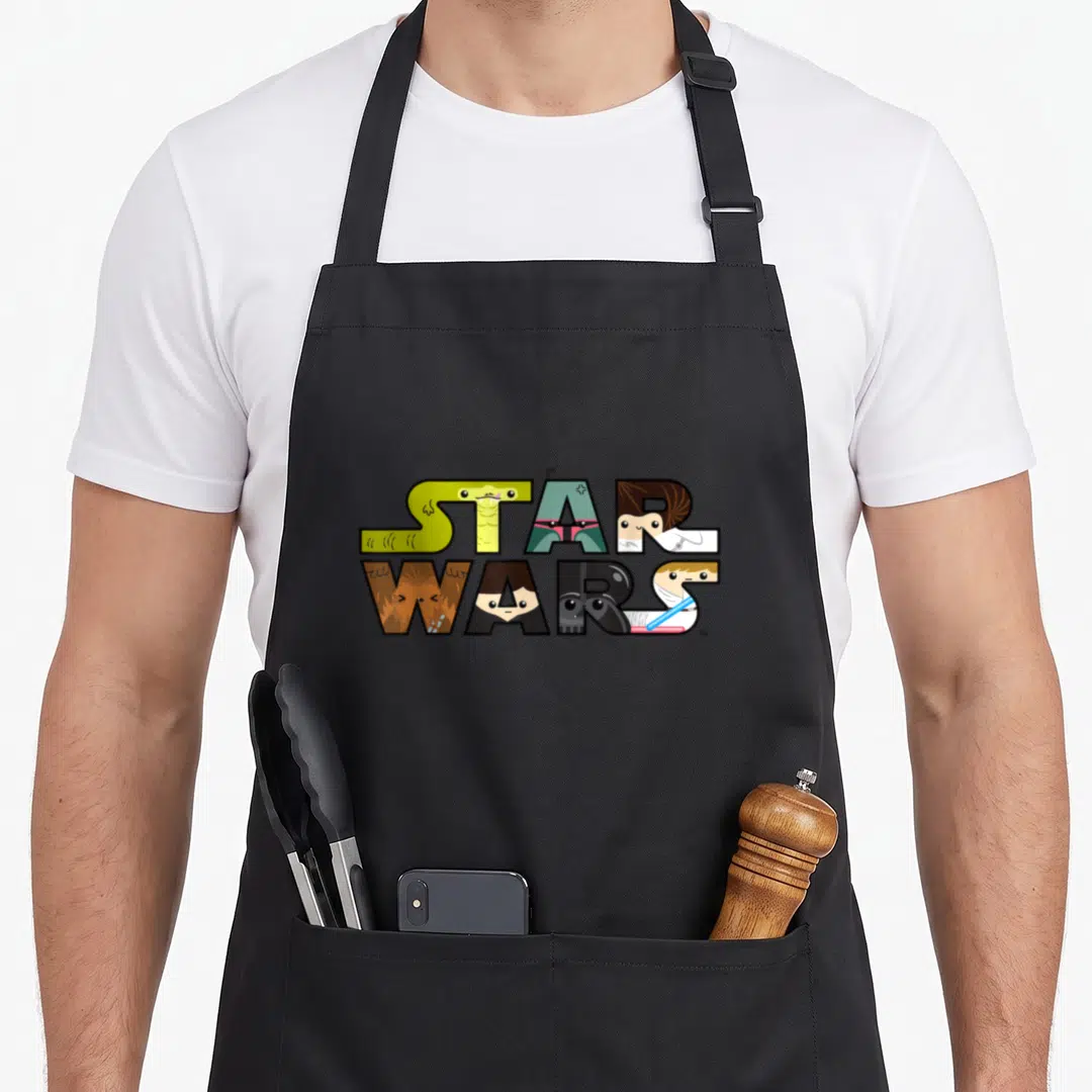 Character kawaii apron - Star Wars text filled with cartoon character faces in green, teal, brown, black, and white — shown on a black cotton canvas apron against a white background