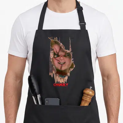 Play door apron - Chucky doll face through torn paper with red CHUCKY text below — shown on a black cotton canvas apron against a white background