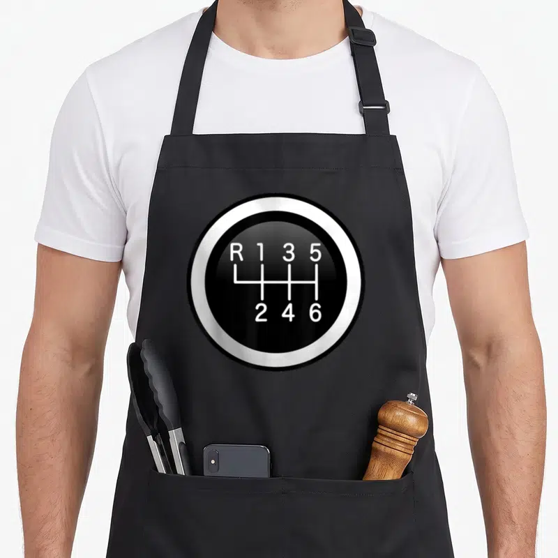 Speed auto apron - Black circle gear shift knob with white R 1 3 5 2 4 6 numbering pattern — shown on a black cotton canvas apron against a white background