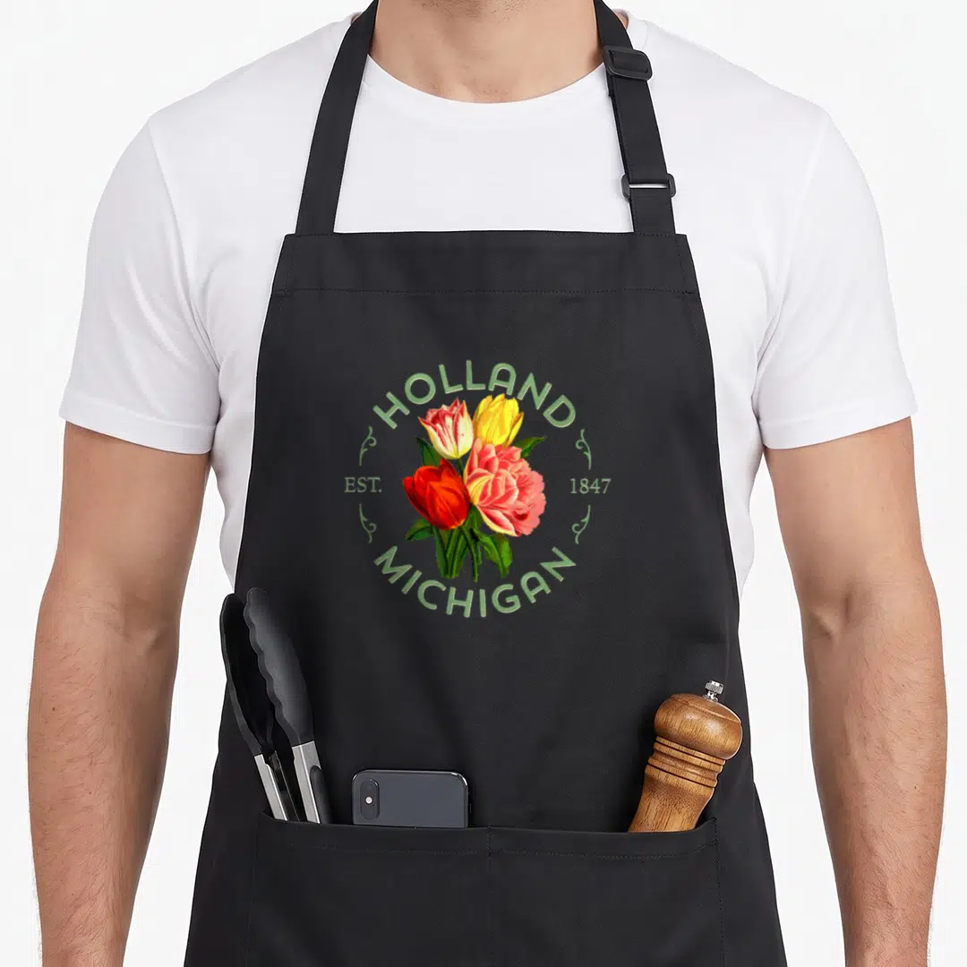Michigan tulip apron - Three red yellow and pink tulips framed by green HOLLAND MICHIGAN EST 1847 text — shown on a black cotton canvas apron against a white background