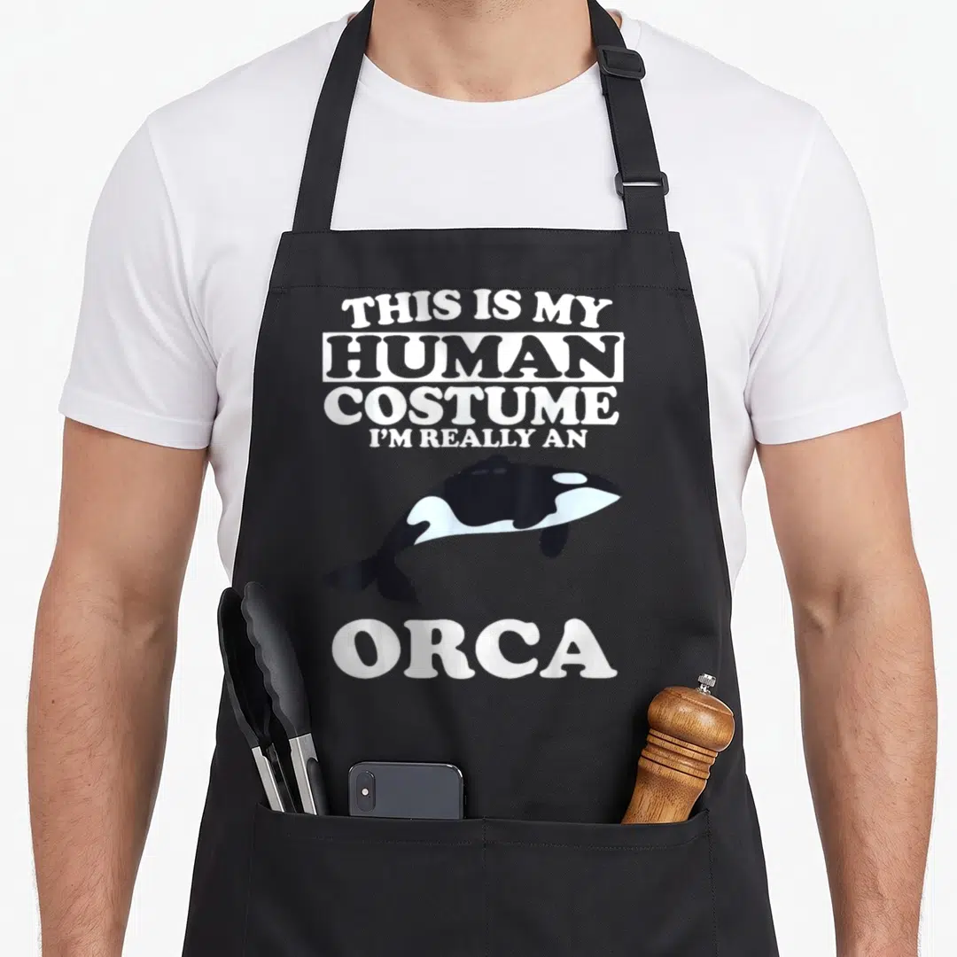 Human orca dad apron - Cartoon orca with text THIS IS MY HUMAN COSTUME I'M REALLY AN ORCA — shown on a black cotton canvas apron against a white background