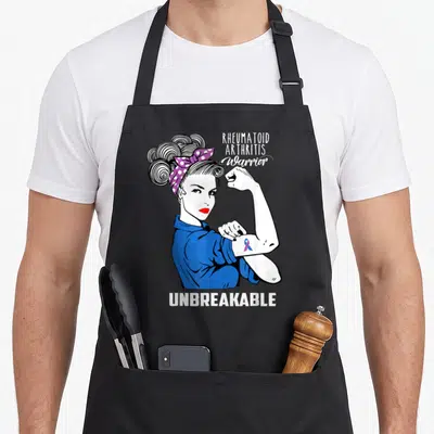 Unbreakable rheumatoid arthritis awareness apron - Cartoon woman in blue shirt flexing arm with purple polka dot scarf — shown on a black cotton canvas apron against a white background