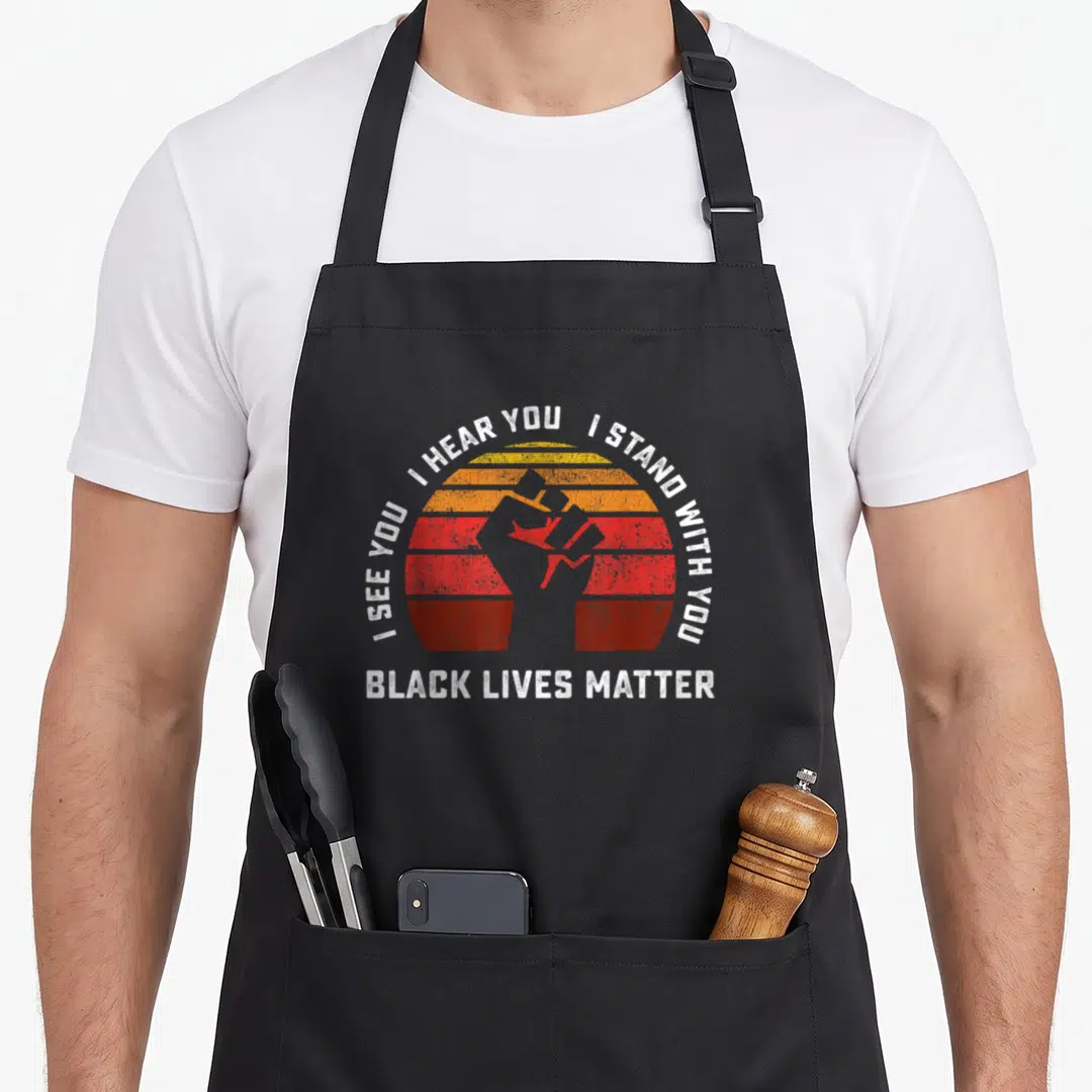 See hear apron - Raised fist silhouette on red-orange-yellow striped circle with white text I See You I Hear You I — shown on a black cotton canvas apron against a white background
