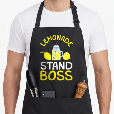 Stand lemonade apron - Cartoon mason jar with lemon slices and stacked LEMONADE STAND BOSS text — shown on a black cotton canvas apron against a white background