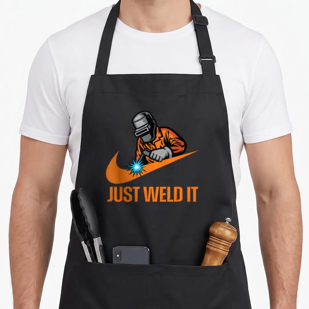 Weld welder apron - Cartoon welder making blue spark on orange swoosh with JUST WELD IT text below — shown on a black cotton canvas apron against a white background
