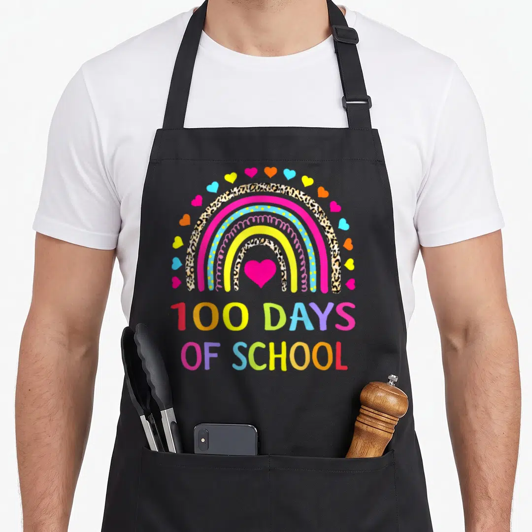 Funny school rainbow apron - Cartoon rainbow with pink heart and leopard print arches over 100 Days of School text — shown on a black cotton canvas apron against a white background