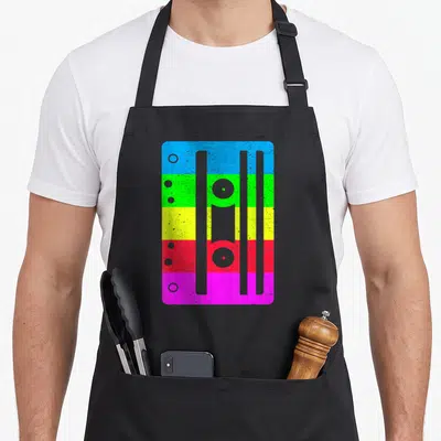 Cassette 80s apron - Retro cassette tape with five horizontal colored bands in blue, green, yellow, red, and pink — shown on a black cotton canvas apron against a white background