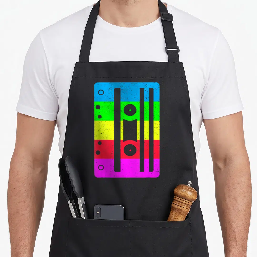 Cassette 80s apron - Retro cassette tape with five horizontal colored bands in blue, green, yellow, red, and pink — shown on a black cotton canvas apron against a white background