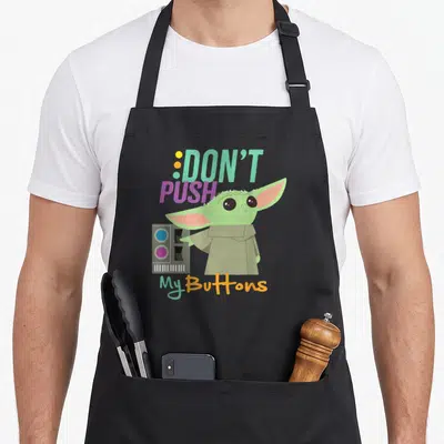 Push wars new apron - Cartoon Grogu reaching for grey panel with DON'T PUSH My Buttons text in green, pink, orange, grey — shown on a black cotton canvas apron against a white background