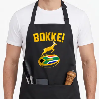 Springbok south african apron - Yellow BOKKE! text over leaping springbok above rugby ball with South African flag — shown on a black cotton canvas apron against a white background