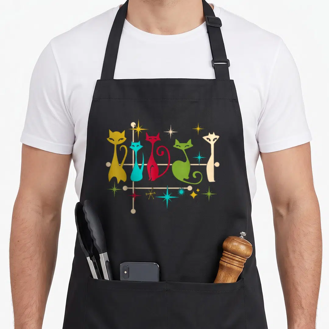 Cat 50s modern apron - Five stylized cats in yellow, blue, red, green, and beige on a beige grid with four-pointed stars — shown on a black cotton canvas apron against a white background