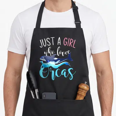 Sea creature animal apron - Just a girl who loves orcas text with jumping cartoon whale over blue waves — shown on a black cotton canvas apron against a white background