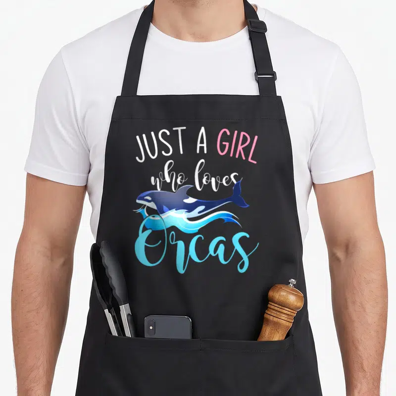 Sea creature animal apron - Just a girl who loves orcas text with jumping cartoon whale over blue waves — shown on a black cotton canvas apron against a white background