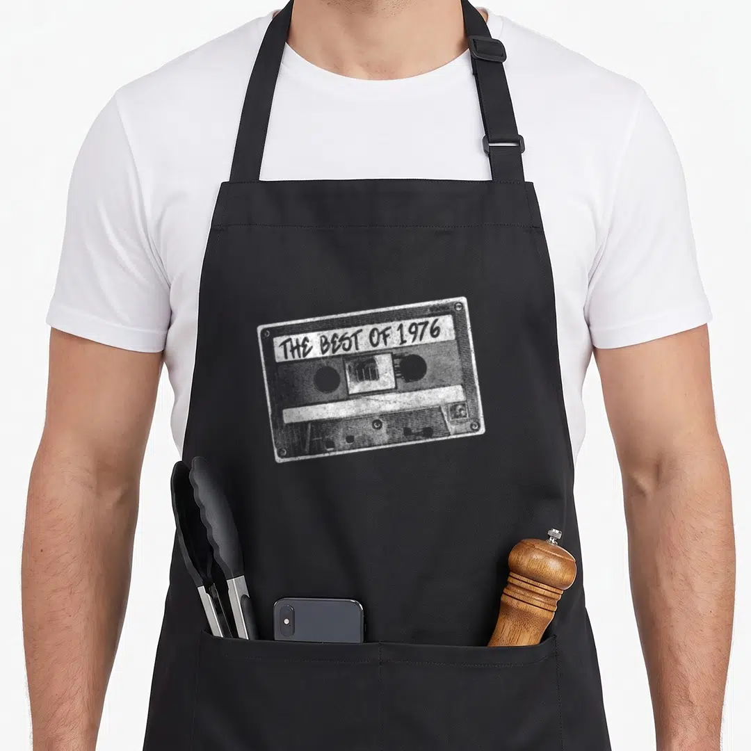 Cassette tape birthday apron - White distressed cassette tape with text The Best of 1976 on the label — shown on a black cotton canvas apron against a white background
