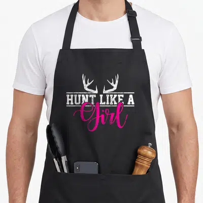 Funny hunting apron - White antlers over HUNT LIKE A text with pink Girl script in distressed style — shown on a black cotton canvas apron against a white background