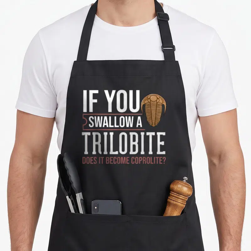 Future archaeologist apron - Trilobite illustration next to stacked text asking about swallowing fossils becoming — shown on a black cotton canvas apron against a white background