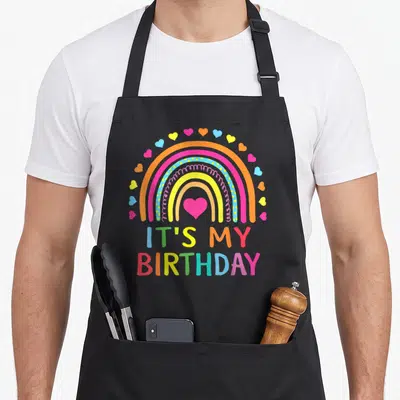 Birthday rainbow apron - Cartoon rainbow with pink heart and scattered hearts above IT'S MY BIRTHDAY text — shown on a black cotton canvas apron against a white background