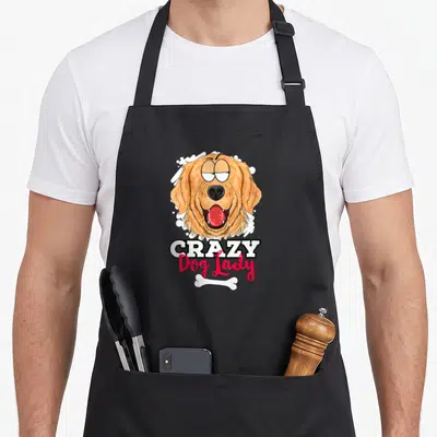 Present owner dog apron - Cartoon golden retriever head above CRAZY Dog Lady text and white bone on splatter background — shown on a black cotton canvas apron against a white background