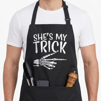 Boyfriend halloween apron - White distressed SHE'S MY TRICK text above a pointing skeleton hand — shown on a black cotton canvas apron against a white background